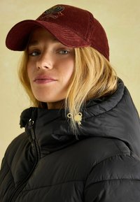 Burgundijas krāsas korduroja cepure ar izliekto malu. Nēsāta ar melnu pūkaino jaku ar augstu apkakli un zelta akcentiem.