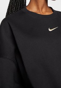 Svart bomullströja med rund hals, sänkt axel och en liten guldfärgad Nike-logga på bröstet. Mjuk textur och enkelt design.