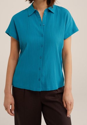 Kvinde iført en kortærmet, knappet grønblå bluse med en subtil krølet tekstur og mørkebrune bukser, stående med armene afslappet ned langs siderne.