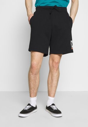 Mężczyzna w czarnych szortach zaciąganych na sznurek z bocznymi kieszeniami, białych skarpetkach i czarnych niskich trampkach na jednolitym jasnym tle.