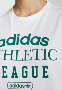 Valkoinen huppari, jossa on vihreä teksti, joka lukee "adidas ATHLETIC LEAGUE" ja logo, jossa on kolme raitaa, pyöreä kaula-aukko ja pehmeä tekstuuri.