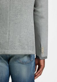 Grijze, gemêleerde blazer met vier knopen aan de mouwen, gecombineerd met blauwe spijkerbroeken. De blazer heeft een eenvoudig ontwerp en een nette snit.
