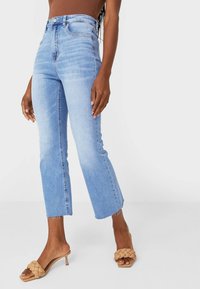 Lichtgewassen, high-waisted jeans met een uitlopende pijp en een onafgewerkte zoom. Gecombineerd met tankleurige, gevlochten slippers. Gladde denimtextuur.