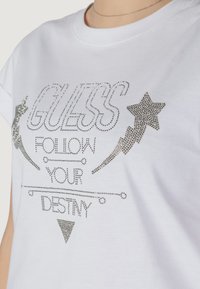 T-shirt bianco in cotone con lettering in strass argentati che legge "GUESS FOLLOW YOUR DESTINY", con accenti di stelle e fulmini.