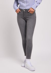 Jeans ajustados grises con un lavado medio, cinco bolsillos y dobladillos deshilachados, combinados con zapatillas blancas y una camisa de botones a rayas.