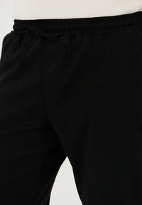 Schwarze Hose aus leichtem Stoff mit elastischem Bund, verstellbarem Kordelzug und Seitentaschen. Glatte Textur mit matter Oberfläche.