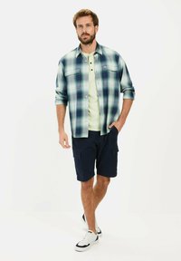 Casual outfit med en grøn og blåternet skjorte, lysegrøn t-shirt, navy cargo shorts og hvide sneakers med sorte detaljer.