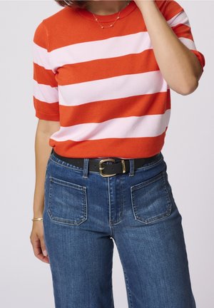 Mujer con camiseta de manga corta con rayas horizontales naranjas y blancas, jeans azules con bolsillos delanteros con parche y cinturón negro con hebilla dorada.