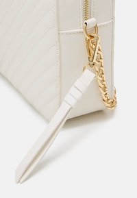 Bílá prošívaná kabelka s texturovaným povrchem, vybavená zlatým řetízkovým popruhem a zipovým uzávěrem. Doplněná o odnímatelný popruh.