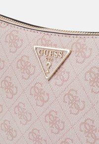 Ljust rosa texturerad handväska med ett diamantmönster, med en guldfärgad triangel-logoplåt som visar "GUESS USA." Dragkedjestängning på toppen.