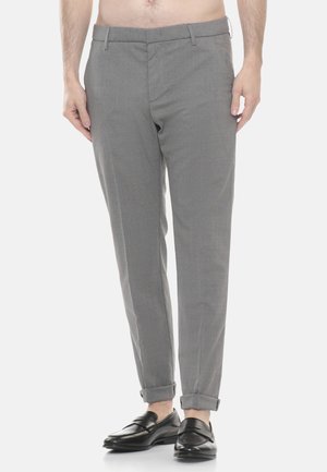 Uomo che indossa pantaloni grigi aderenti con orli risvoltati e mocassini neri in pelle, in piedi contro uno sfondo chiaro e uniforme.