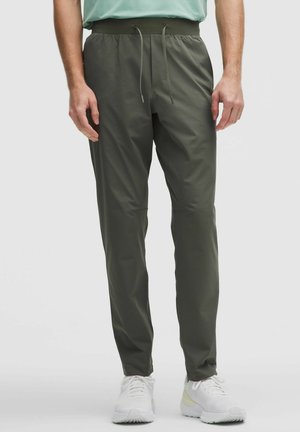 Homme portant un pantalon à cordon de serrage vert olive et des baskets blanches sur un fond clair uni.