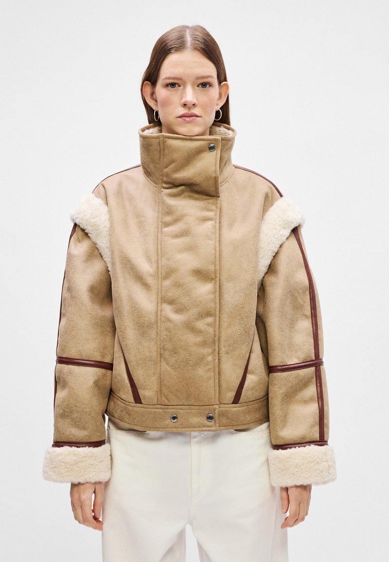 Braune Lammfelljacke mit hohem Kragen, plüschigen weißen Manschetten und kastanienbraunen Akzenten. Verfügt über Druckknopfverschlüsse und eine kastige Form.