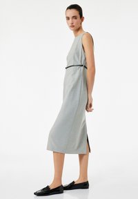 Kobieta stojąca w szarej, bezrękawnikowej midi sukience z wąskim czarnym paskiem i czarnymi loafersami na tle jednolitego białego tła.