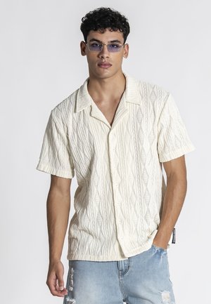 Ung mand iført en tekstureret off-white kortærmet knappeskjorte, lyseblå revnede jeans og tonede rektangulære briller, stående med den ene hånd i lommen.