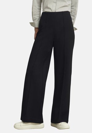 Person trägt hoch taillierte schwarze weit geschnittene Hose mit einem weiß-schwarz gestreiften Langarmshirt und weißen Turnschuhen vor einfachem Hintergrund.