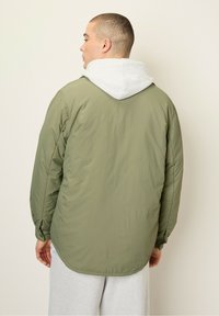 Olivengrøn pufferjakke med en grå hætte. Har elastiske manchetter og en buet kant. Glat tekstur og letvægtsdesign.