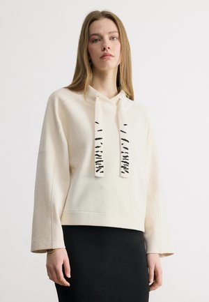 Jeune femme aux longs cheveux portant un sweat à capuche crème avec des cordons de serrage noirs à motifs et une jupe noire, sur un fond clair uni.