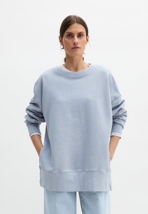 Kvinde med mørkt hår samlet bagud iført en løs lyseblå sweatshirt og matchende bukser, stående med hænderne i lommerne mod en hvid baggrund.