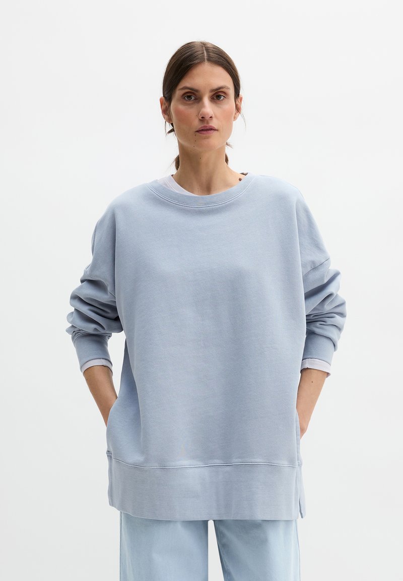 Γυναίκα με σκούρα μαλλιά δεμένα πίσω, που φοράει άνετο ανοιχτό μπλε φούτερ και ταιριαστό παντελόνι, όρθια με τα χέρια στις τσέπες μπροστά σε λευκό φόντο.