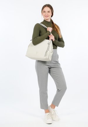 Sac à main texturé beige avec une bandoulière, doté d'un détail à frange et d'un logo. Le modèle porte un haut côtelé vert et un pantalon gris ajusté.