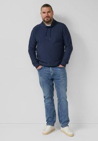 Marineblauer Pullover mit hohem Kragen, Kordelzug und gerippten Bündchen, kombiniert mit hellblauen Denim-Jeans und weißen Turnschuhen.