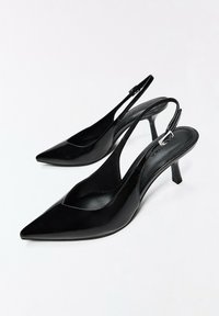 Escarpins slingback à talons kitten en cuir verni noir avec bouts pointus et brides réglables à boucle sur fond blanc.