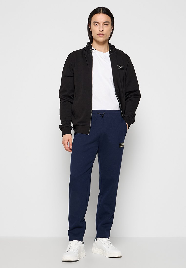 Schwarze Zip-Hoodie mit hohem Kragen, darunter ein weißes T-Shirt und marineblaue Jogginghosen mit einem dezenten Logo, kombiniert mit weißen Turnschuhen.