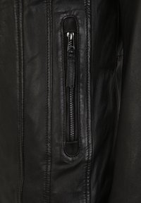 Černá kožená bunda s postranní kapsou na zip. Má hladkou texturu, štíhlý design a kontrastní stehy podél švů.