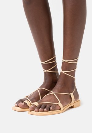 Flache Sandalen mit einer beigen Gummisohle, die ein gekreuztes Riemendesign aus dünnem, strukturiertem Material aufweisen. Offen und mit minimalistischem Stil.