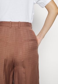 Personne portant un pantalon marron texturé taille haute avec des poches fendues à l'arrière et une chemise blanche à manches courtes, la main posée sur la hanche.