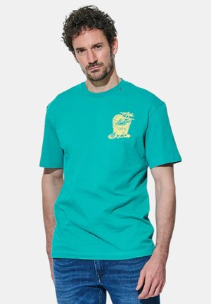 Uomo con capelli ricci che indossa una maglietta turchese con una grafica gialla di una bevanda tropicale sul petto e jeans blu su uno sfondo semplice.