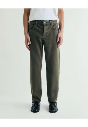 Hombre con pantalones de corte relajado de color verde oscuro, zapatos negros con cordones y camisa blanca metida, de pie contra un fondo claro y liso.