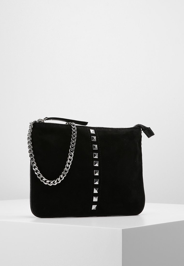 Clutch rectangular negro de ante con tachuelas plateadas verticales y una correa de cadena plateada, exhibido sobre un pedestal blanco contra un fondo liso.