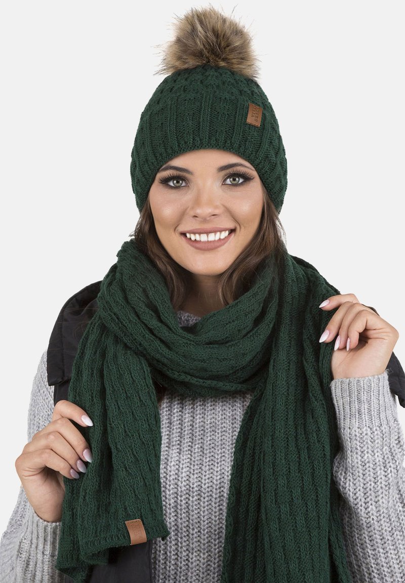Gorro de lana verde con un pompón de piel sintética, combinado con una bufanda texturizada a juego. Ambas piezas presentan un diseño acanalado y un acento de etiqueta de cuero.