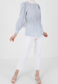 Vaaleansininen paita, jossa on elastinen vyötärö, puffihihat ja tekstuuripinta, yhdistettynä valkoisiin suoralahkeisiin housuihin ja valkoisiin hihnatossuihin.