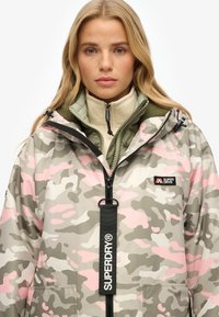Veste camouflage dans des tons gris, blanc et rose avec fermeture éclair avant, capuche ajustable et étiquette logo Superdry. Présente un design superposé.