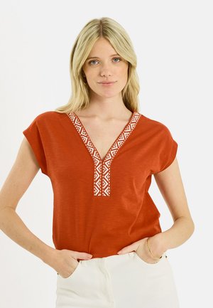 Mujer rubia con una blusa de cuello en V color óxido con bordados geométricos blancos, con las manos en los bolsillos, de pie contra un fondo blanco.