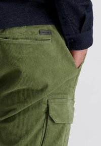 De hand van een man in de zak van groene corduroybroek met een klein zakflapje en een "Pierre Cardin" label, gekleed in een donkerblauwe lange mouwen shirt.