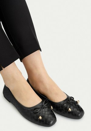 Pies con bailarinas tejidas negras con pequeños cascabeles dorados en los lazos delanteros, combinadas con pantalones negros recortados sobre un fondo liso.