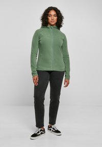 Green fleece jacket with a zip closure, a high collar, and side pockets, paired with black jeans and black sneakers. Smooth texture.