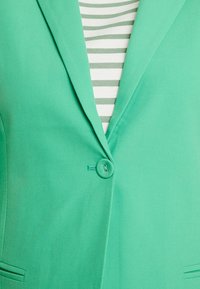 Blazer vert avec une fermeture à un seul bouton, tissu lisse et revers crantés, superposé à une chemise blanche et verte à rayures.
