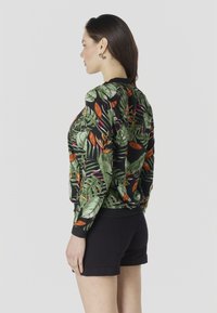 Vrouw met donker haar die zwarte shorts en een lange mouwen top met tropische bladerenprint draagt, staand in profiel tegen een witte achtergrond.