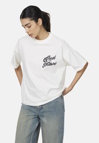 Donna in una maglietta bianca larga con la scritta "Good for Nothing" e jeans azzurri chiari, che guarda in basso con le mani appoggiate sui fianchi.
