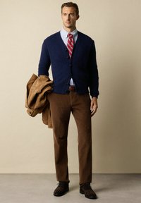 Cardigan in lana navy con chiusura a bottoni, camicia a righe, cravatta rossa, pantaloni in velluto a coste marroni e scarpe marrone scuro, mentre tiene un giubbotto in suede beige.