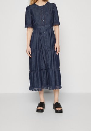 Femme portant une robe midi bleu foncé avec boutons, manches ballon, bordure en dentelle et sandales plateformes noires, debout sur un sol gris.