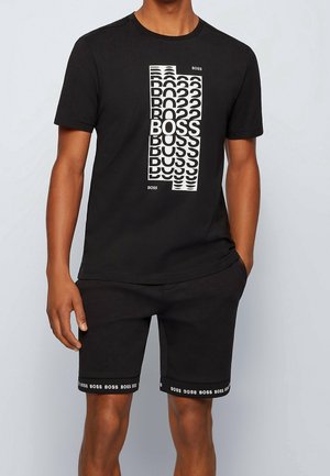 Homme portant un ensemble T-shirt et shorts noirs avec des motifs répétés du logo « BOSS » sur le devant et l'ourlet des shorts.