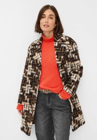 Wollen blend jas in een bruine, witte en zwarte zigzagpatroon, gecombineerd met een feloranje sweater en donkergrijze jeans. Luipaardprintriem zichtbaar.