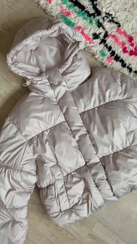 Lysegrå boblejakke med hette og to frontlommer, lagt flatt på tregulv ved siden av et hvitt teppe med svarte, rosa og grønne mønstre.