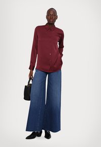 Burgunderfarget satengbluse med krage, knappefront og lange ermer, kombinert med vide denimjeans. Modellen holder en svart håndveske.